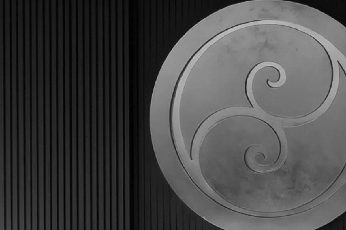 Black-and-white close view of the entrance wall: the circular monogram engraved in metal, set into a wall of fine dark vertical slats.